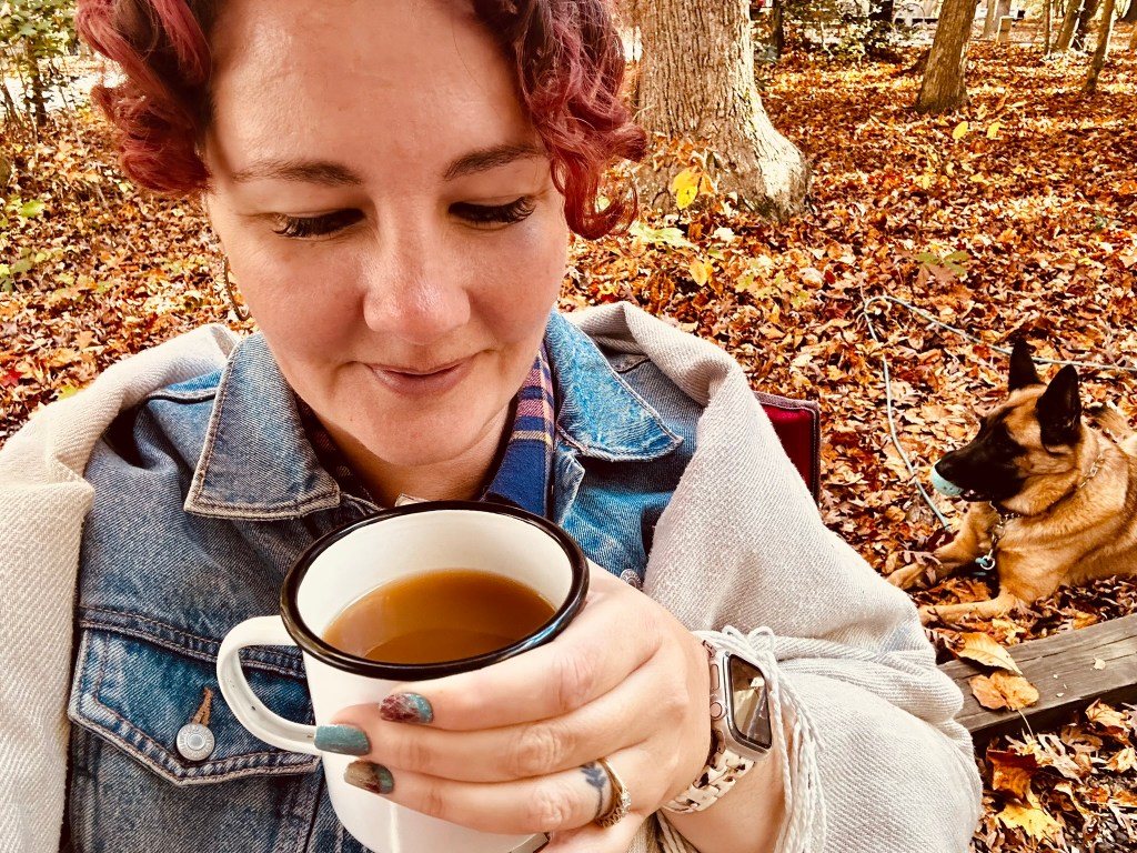 A person with curly hair holding a cup of coffee, sitting amongst a bed of autumn leaves with a dog in the background.